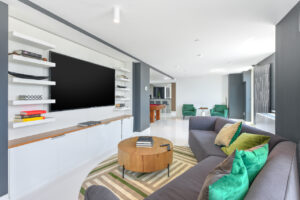 Interior Community Room, gray walls, large sectional couch, wood coffee table, white built tv console, contemporary rug, large tv, green lounge chairs with an end table between, pool table in other room.