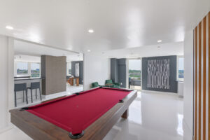 Interior Community Room, red pool table, contemporary art on walls, gray and white walls, bar area in other room, tv area in other room, green lounge chairs near pool table, white floor.