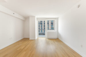 Interior Unit Living Room, door to balcony with view of nearby building, wood floors, white walls.